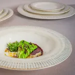Bonna Galata Round Coupe Bowl in use, holding a plated dish of grilled vegetables in a matte off-white ceramic vessel with a flared ribbed rim.