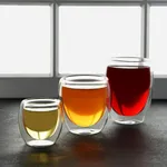 Brew Infusion Double Wall Glass 220ml 2pc Set in use, three tumblers with pale yellow, amber, and red liquids on dark stone.