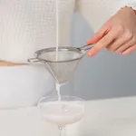 D-Still Fine Strainer 100mm Stainless Steel in use, pouring liquid through conical mesh into a coupe glass
