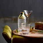 D-Still Royal Cocktail Shaker in a lifestyle scene with gold-toned cap, crystal body, and glassware on a wooden table