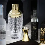 Close-up of the D-Still Royal Cocktail Shaker with gold cap and textured glass body, displayed with a jigger and bottles.