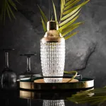 Close-up of the D-Still Royal Cocktail Shaker with a clear diamond-cut glass body and gold-toned metal cap resting on a wooden coaster.