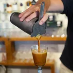 D-Still Hawthorn Strainer in use, pouring amber liquid from a shaker into a glass, featuring a perforated disc and slender handle.