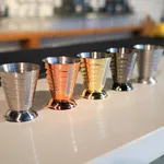Five D-Still Multi Level Jigger 75ml Black tools in copper, gold, and steel finishes, showing engraved volume markings on a diagonal row.