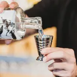 D-Still Multi Level Jigger 75ml Stainless Steel in use, catching a pour from a bottle with visible horizontal ridges on the tapered body.