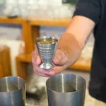 D-Still 75ml vintage stainless steel jigger in use, held by a hand with visible measurement markings and a flared footed base.