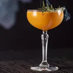 Libbey Nick and Nora glass in use, filled with an orange cocktail and garnished with rosemary and cucumber