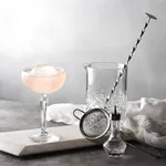 Libbey Onis Speakeasy Gin & Tonic glassware in use, featuring a faceted crystal pitcher with a bar spoon and a coupe holding a pink cocktail with an ice sphere.