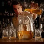 Libbey Tulip Bowl & Spoon-4lt in use, pouring amber liquid from a pitcher into the textured clear glass carafe.