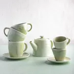 Close-up of matte sage-green Moda Porcelain Lush Coffee and Tea Cups and lidded teapot stacked on saucers