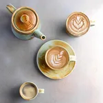 Moda Porcelain Nourish Coffee / Tea Cup in use with latte art, featuring mottled cream and rust-brown glaze with a pale blue-green rim.