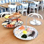 Chef Inox two-tier seafood stand in use on a table, holding raw oysters and grilled fish on stainless steel tiers.