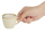 Olympia Kiln Sandstone Espresso Cup in use, held by a hand, showing pale cream mottled ceramic body and brown glaze drip.