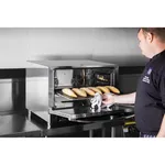 Apuro convection oven in use with door open, revealing golden baguettes on a wire rack as a chef slides the tray out.