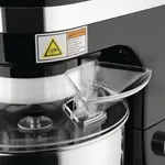 Close-up of the black Apuro Planetary Mixer showing the clear plastic ingredient pourer on the stainless steel bowl and a white safety warning sticker.