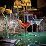 Luigi Bormioli Nick & Nora glass in use with amber cocktail and orange garnish, displayed with other stemware on a green table