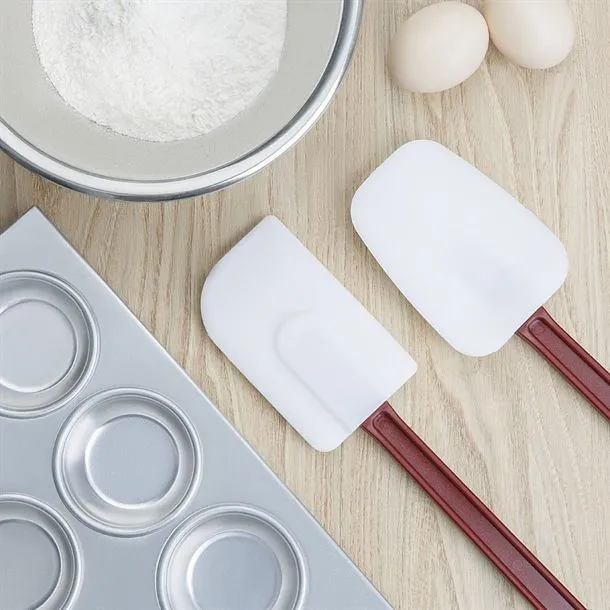 Close-up of two white silicone spoonulas with reddish-brown handles in a baking scene with flour and eggs