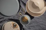 Luzerne Round Plate 280mm Ø in an overhead view of a speckled stoneware set with a grey plate and beige bowl on dark linen.