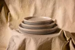 Luzerne Stackable Round Plate 200mm Ø, side view of four speckled beige and grey ceramic plates stacked on linen.