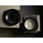 Dudson Round Coupe Bowl overhead view showing matte black speckled exterior and light beige interior on stacked stone slabs
