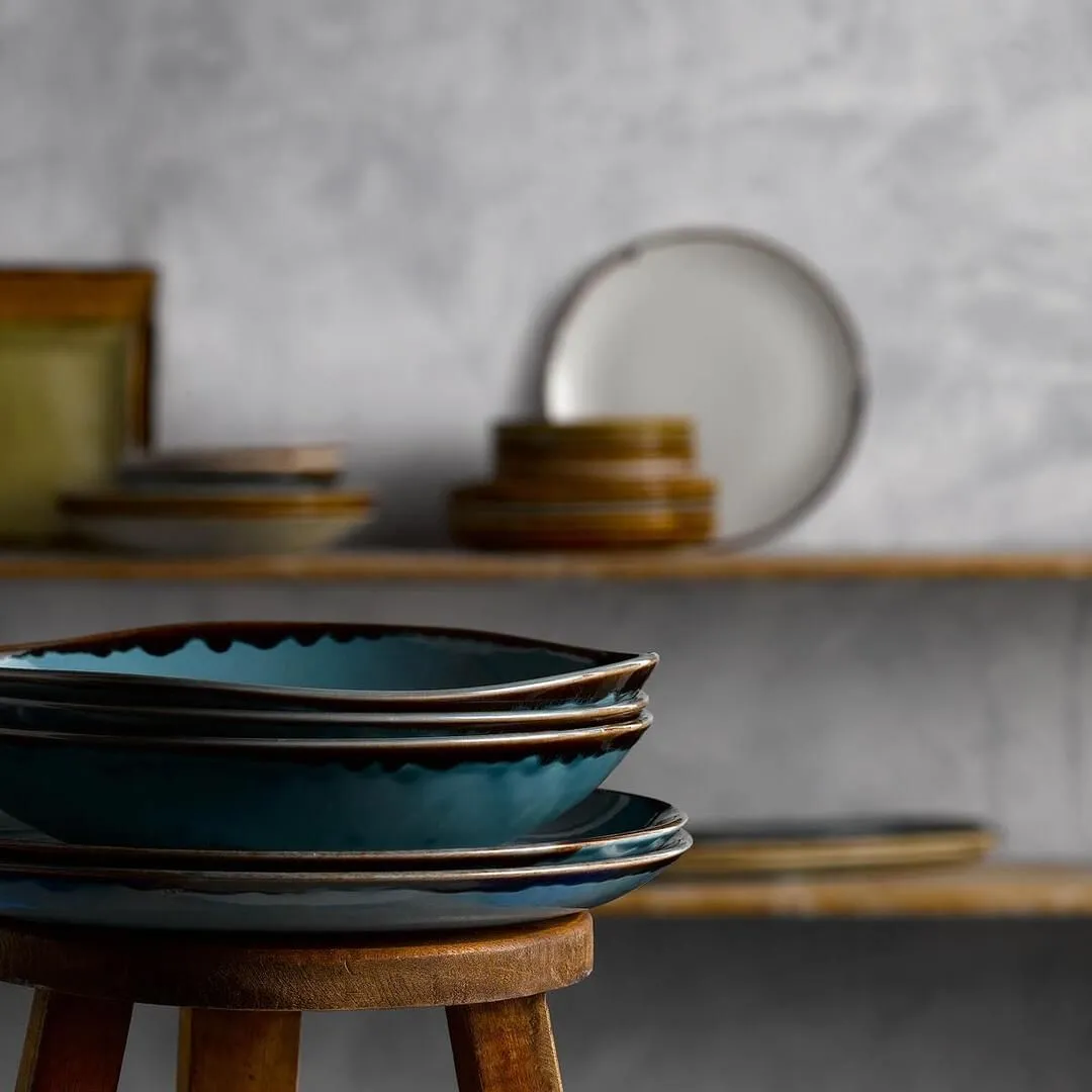 Teal-glazed ceramic plates and bowls with dark brown rims stacked on a wooden stool in a mixed tableware set