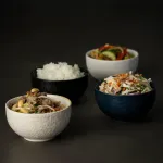 Luzerne Round Plate-Flat, 180mm Ø, shown in an in-use scene with four textured ceramic bowls filled with rice, stir-fry, salad, and noodles.
