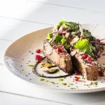 Close-up of the Luzerne Round Bowl 190mm 1400ml, a speckled beige ceramic vessel holding a plated fish dish with fresh salad.