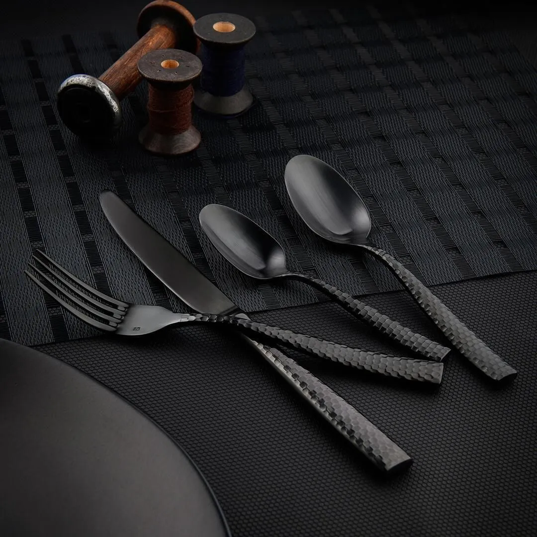 Fortessa Lucca Faceted Dessert Spoon in matte black with textured scale-pattern handles, shown in a close-up with other cutlery on a woven placemat.