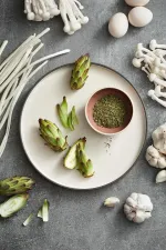 Luzerne Round Stackable Plate in Dusted White with dark grey rim, shown in use holding artichokes and herbs.