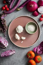 Luzerne Smokey Plum round plate in use, holding garlic and salt, surrounded by vegetables on a stone surface.